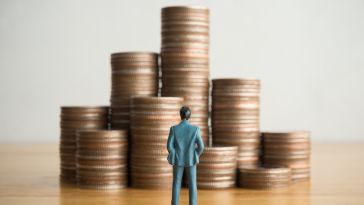 A miniature man in a business suit stands on a table looking at nine stacks of pennies.