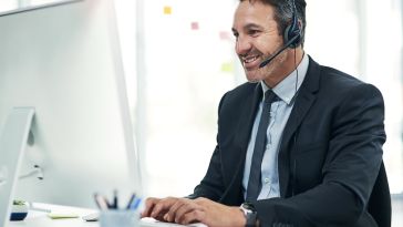 A smiling male professional wearing a business suit and a headset sits at his desk while typing on a computer keyboard in a bright office setting.
