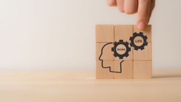 A photo of a hand placing wooden blocks with images of gears that say work and life next to a human head profile to show the idea of work-life balance and employee wellbeing. 