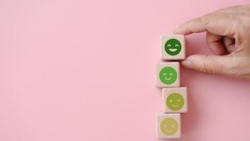 A photo of emoticon faces on a stack of wooden blocks and a handing holding the top one to show the idea of positive employee wellbeing. 