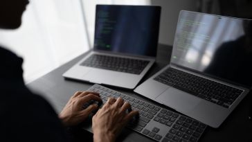 A person types on a full-sized external keyboard at a dark desk positioned in front of two open laptops displaying lines of green code on their screens.