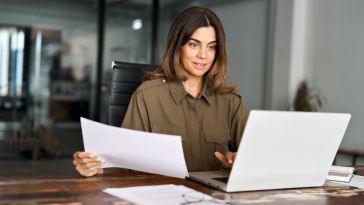 A professional woman sits at a wooden office desk, holding a document and smiling slightly as she looks at her open laptop screen.