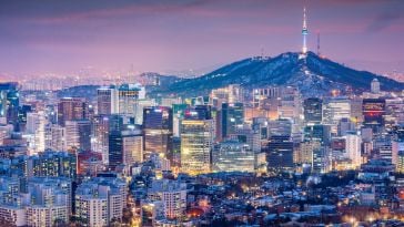A view of the Seoul skyline at night featuring dozens of lit up high-rise buildings in the foreground and mountains in the background.