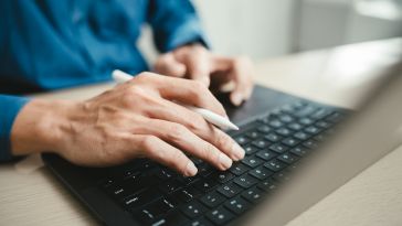 A close-up shot of a person wearing a blue shirt typing on a laptop keyboard and using the trackpad while holding a white stylus.
