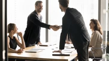 Two businessmen in suits smile as they shake hands across a conference table in a modern office, surrounded by seated colleagues observing the meeting.