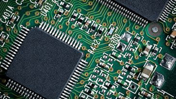A close-up view of a green printed circuit board featuring an integrated circuit microchip surrounded by intricate copper traces, resistors and various surface-mounted electronic components.