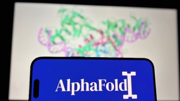 A mobile phone with the AlphaFold logo displayed on it, with an image of a protein structure on the wall behind it.