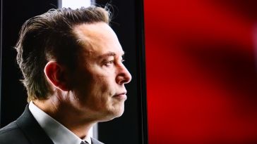 A side-shot of Elon Musk, with a black and red background behind him.