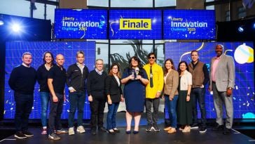 Rashmi Gehani poses for a photo while holding a trophy onstage during the 2025 Liberty Innovation Challenge alongside several company leaders
