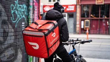 A DoorDash driver sits on a bike next to a wall. 