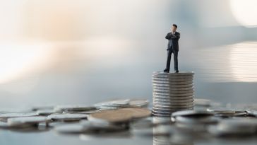 A photo of a miniature figure on a stack of coins to show the idea working in a financial department at a tech company. 