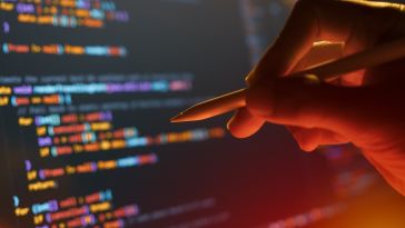 A hand holds a stylus to point directly at colorful lines of computer code displayed on a dark screen under warm lighting.