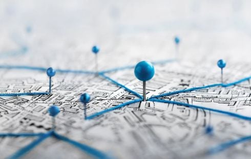 A close-up photograph of a large blue thumbtack stuck in a map, surrounded by blue thread and smaller thumbtacks