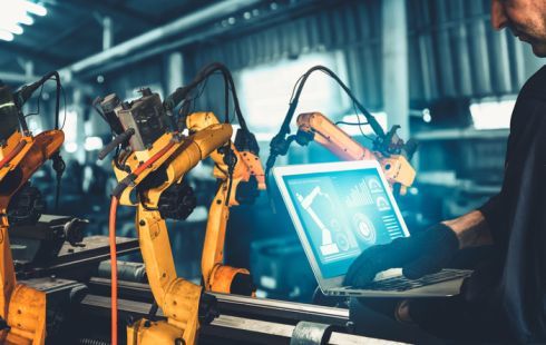 A worker uses a laptop to control a set of robotic arms to represent industrial automationl.