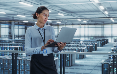 Dark-haired woman wearing a lanyard and looking at a laptop. She stands on a platform looking down at a data center.