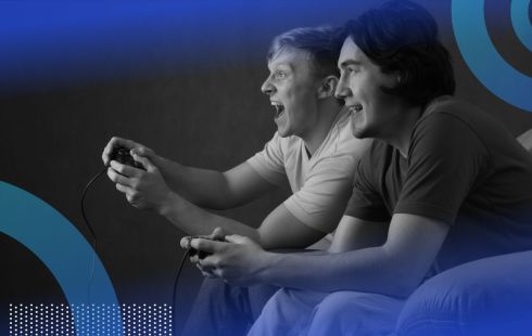 Two young men are holding gaming consoles and looking excitedly at a screen.