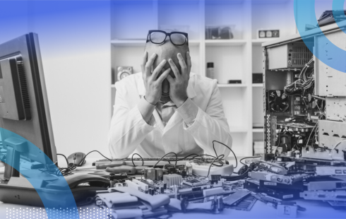 A frustrated man sitting at a desk littered with machine parts with his head in his hands and glasses on top of his head.