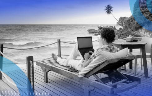 A person is sitting on a lounge chair with a laptop on a patio overlooking an ocean.