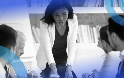 A woman professional standing to lead a meeting while her colleagues sit at the table in front of her.