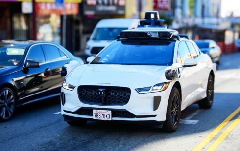 Photo of a white, 4-door sedan fitted with various sensors and driving down a city street.