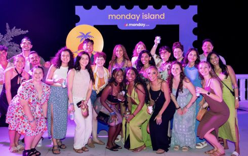 A group of monday employees pose outside by a sign that says &quot;monday island&quot; at night on a beach scene.