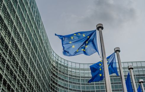 Two European Union flags wave in the wind, with a building surrounding them in the background.