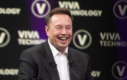 Headshot of Elon Musk laughing in a black suit and white dress shirt.