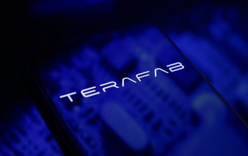 The Terafab logo is displayed on a smartphone screen placed on a reflective surface with blue light projections.