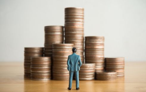 A miniature man in a business suit stands on a table looking at nine stacks of pennies.