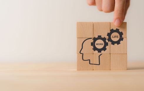 A photo of a hand placing wooden blocks with images of gears that say work and life next to a human head profile to show the idea of work-life balance and employee wellbeing. 