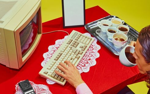 A person typing on the keyboard of a vintage desktop computer while drinking coffee.