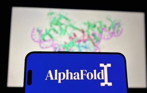 A mobile phone with the AlphaFold logo displayed on it, with an image of a protein structure on the wall behind it.