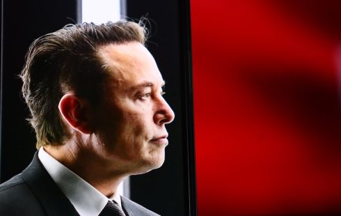 A side-shot of Elon Musk, with a black and red background behind him.