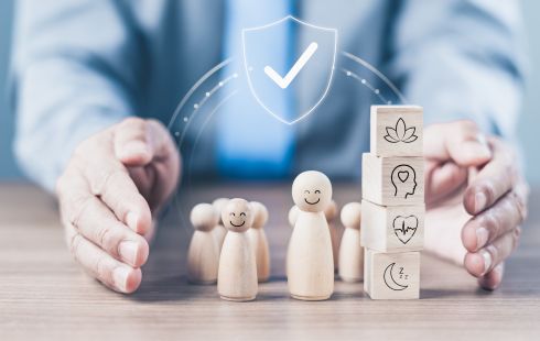 Hands around wooden figures with smiles and blocks stacked next to them with icons like plant, heartbeat pulse and a moon with sleep symbols to show the idea of protecting employee wellbeing. 