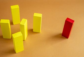 A red wooden block stands apart from a group of yellow wooden blocks, illustrating the marginalization that microaggressions cause in the workplace.