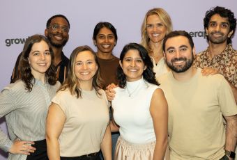 A group of Grow Therapy employees smile in front of a lavender wall printed with the company’s branding.