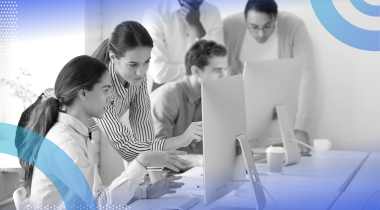 Supervised learning image of a group of professional people working together on a project. A man and a woman are in the foreground. He seeks her help with a problem. She stands next to him pointing at the screen.