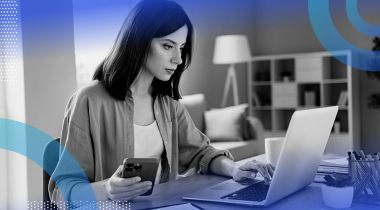 A woman focuses on a laptop screen and uses a smartphone