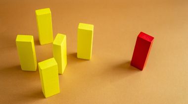 A red wooden block stands apart from a group of yellow wooden blocks, illustrating the marginalization that microaggressions cause in the workplace.
