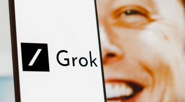 A smartphone with 'Grok' on its screen and the face of Elon Musk peaking behind it.