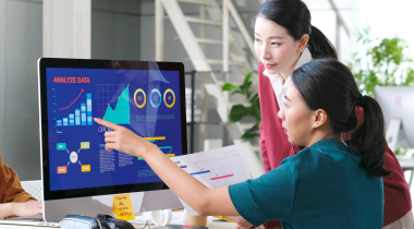 Two women of Asian descent look at graphs on a large desktop screen. The woman in the foreground wears green and points at the screen.