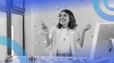 A business woman sitting at a computer desk, smiling with confetti raining down on her.