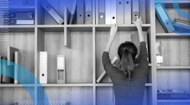 A woman reaching to grab a binder from a bookshelf/cubby hole that’s in disarray.