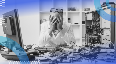 A frustrated man sitting at a desk littered with machine parts with his head in his hands and glasses on top of his head.
