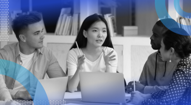 Four young professionals sitting at a desk with laptops having a discussion.