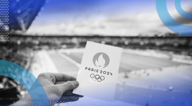 A hand is holding a card that says Paris 2024 in front of an athletic field.