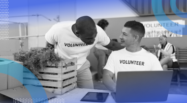 A person wearing a volunteer t-shirt is sitting at a computer and talking to another volunteer.