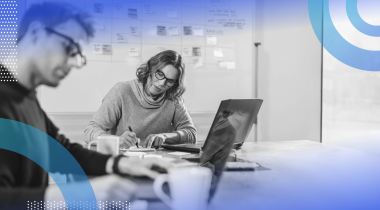 Two people are sitting at a desk working on a project. A whiteboard behind them is filled with sticky notes.