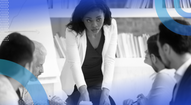 A woman professional standing to lead a meeting while her colleagues sit at the table in front of her.