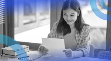 A woman smiling at a piece of paper while sitting next to a notebook, pen and book.
